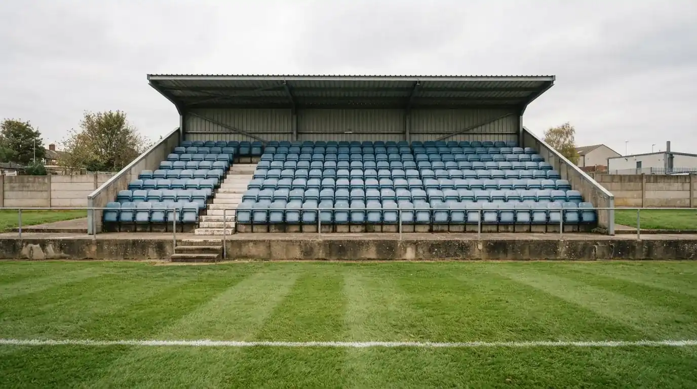 Rzędy pustych plastikowych krzesełek małego stadionu widziane z poziomu murawy pod pochmurnym niebem.